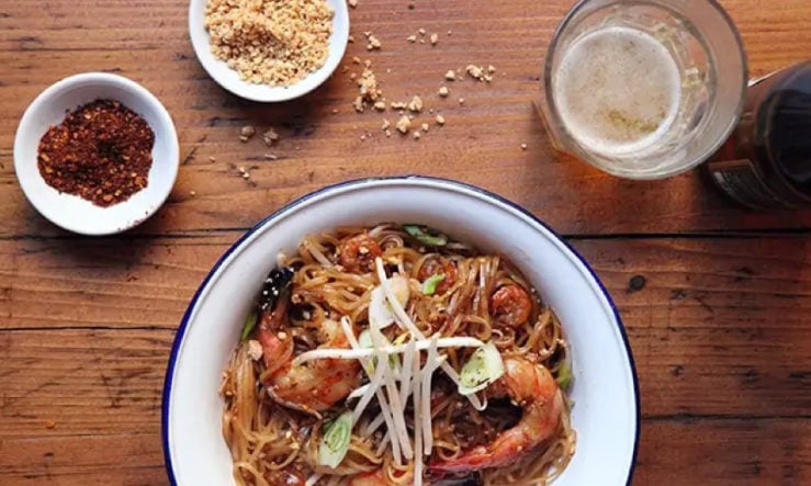 Rosa's Thai Pad Thai served in a white enamal bowl with seasoning and a drink to the side