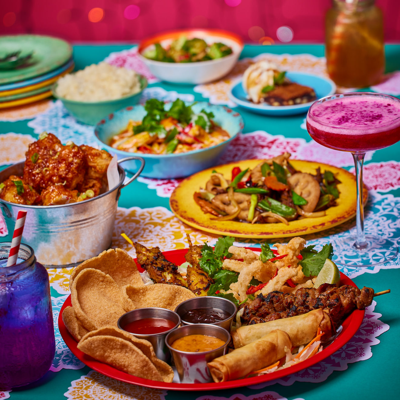 Thai feast food spread with sharing platter in the foreground on a teal table top with colourful doilies, bright pink background with multi coloured fairy lights